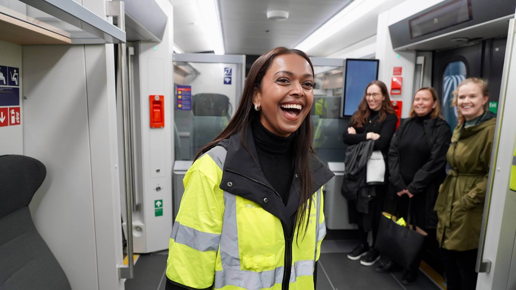 Kamzy Gunaratnam, samferdselsminister, i gul refleksjakke ler i en togvogn med tre andre kvinner stående i bakgrunnen. (Bildebeskrivelsen er laget av en KI-tjeneste)
