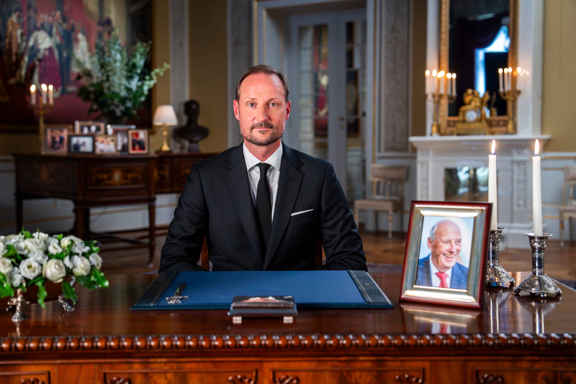 Kong Haakon sitter ved et skrivebord i Slottet, med et portrett av Kong Harald ved siden av. (Bildebeskrivelsen er laget av en KI-tjeneste)