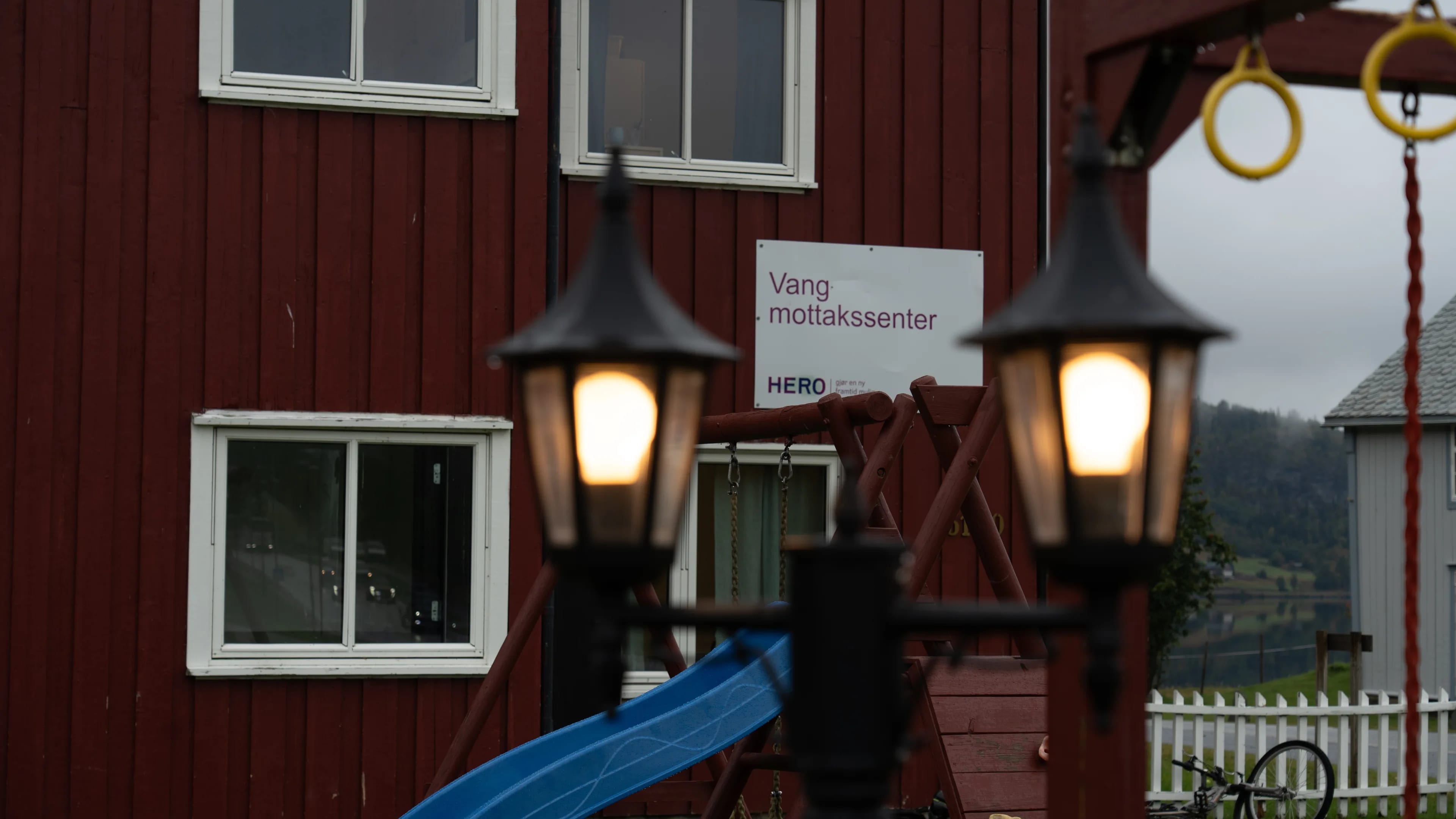 Skilt for Vang mottakssenter på en rød trebygning med lekeapparat i forgrunnen. (Bildebeskrivelsen er laget av en KI-tjeneste)