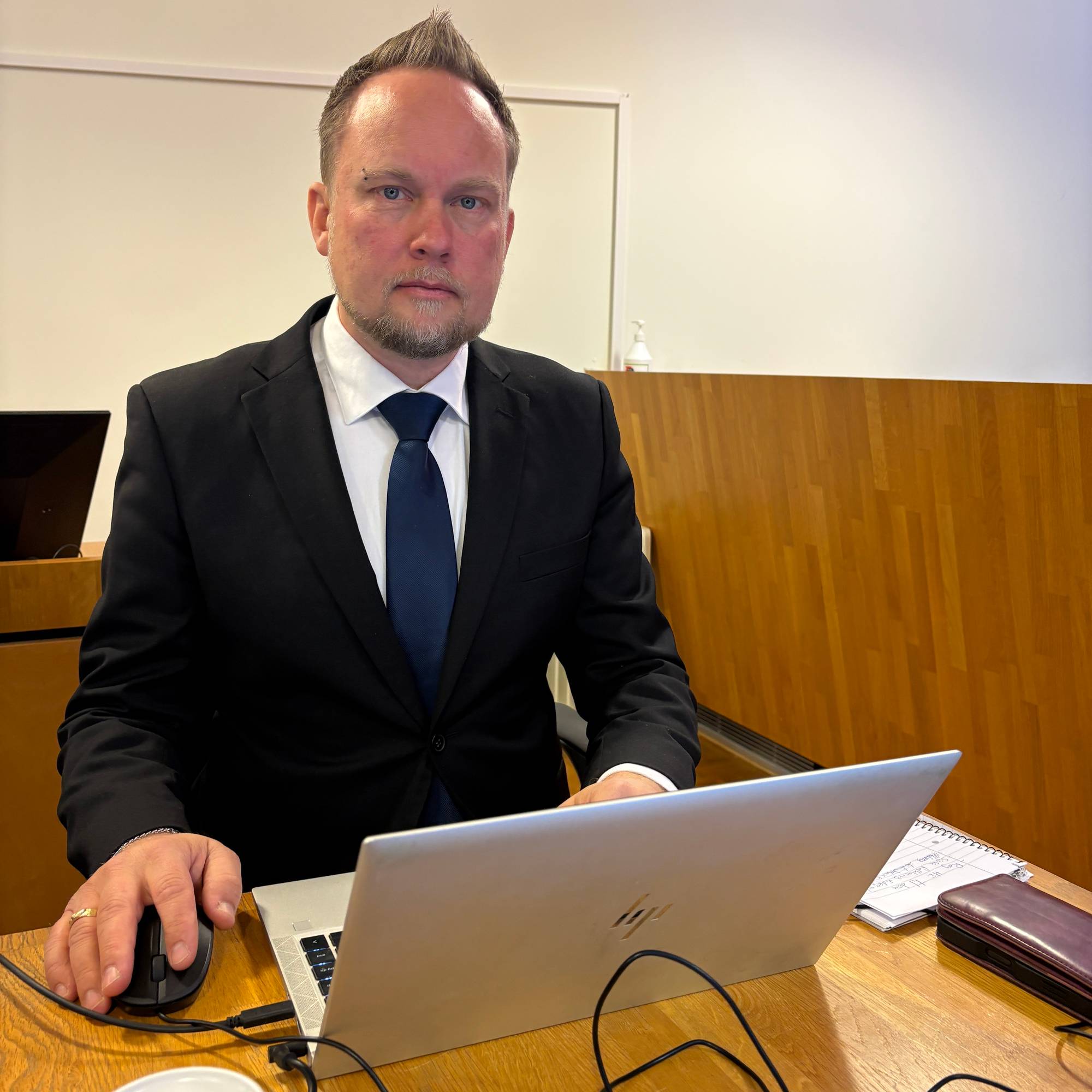 En mann i dress sitter foran en bærbar datamaskin. Han bruker en mus og ser mot kamera. Et glass med vann står på bordet. 