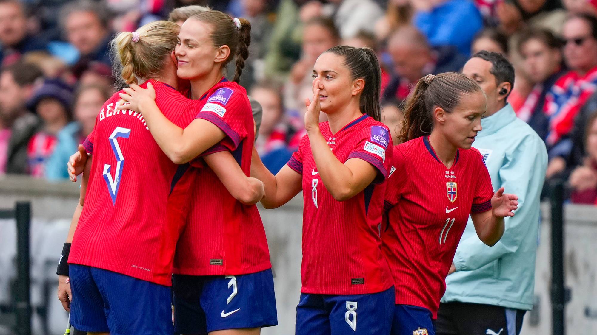 Norske fotballspillere klemmer hverandre på banen. De er iført røde trøyer og blå shorts. En person med lyseblå jakke står bak dem. Tilskuere sitter på tribunen i bakgrunnen. (Bildebeskrivelsen er laget av en KI-tjeneste)