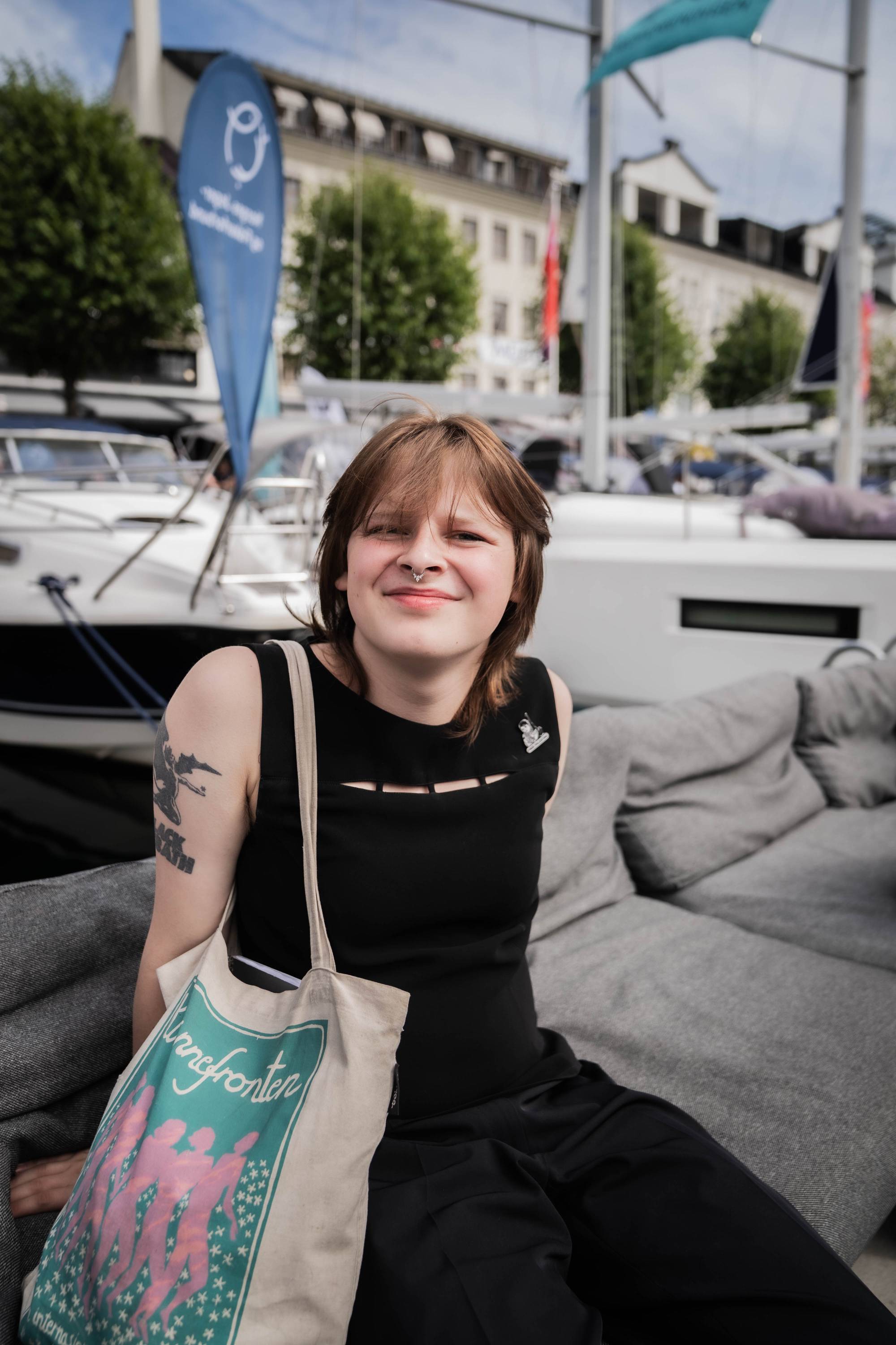 En person sitter i en sofa ved en båthavn og smiler til kamera. I bakgrunnen ligger flere båter. 