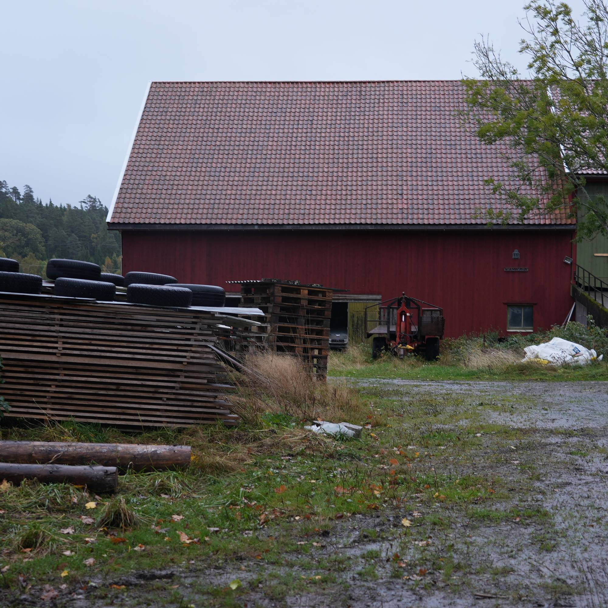 Rød låve på et gårdsstun med trelast, bildekk og landbruksutstyr i forgrunnen. (Bildebeskrivelsen er laget av en KI-tjeneste)