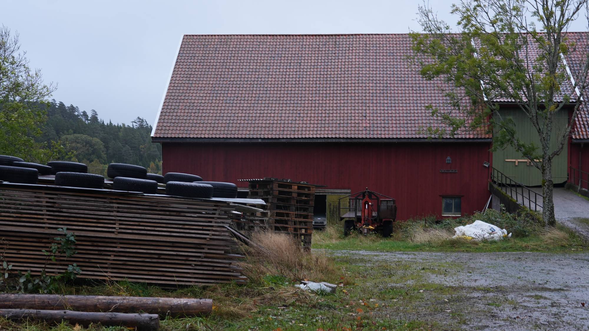 Rød låve på et gårdsstun med trelast, bildekk og landbruksutstyr i forgrunnen. (Bildebeskrivelsen er laget av en KI-tjeneste)