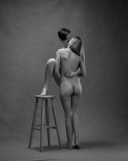 En naken dame står på gulvet og kysser en naken dame med sort hår og mørk neglelakk og fotsålen på en krakk