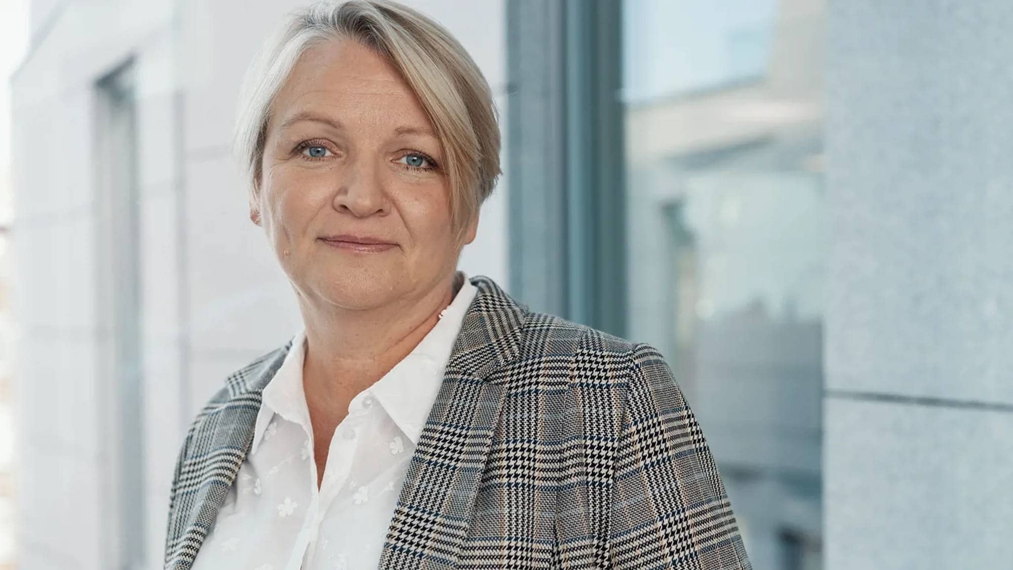En person med kort, lyst hår poserer foran en glassvegg. Personen har på seg en rutete blazer over en hvit skjorte. Omgivelsene er moderne, med synlige geometriske former og lys fra vinduet. Bildeutsnittet fokuserer på overkroppen. (Bildebeskrivelsen er laget av en KI-tjeneste)