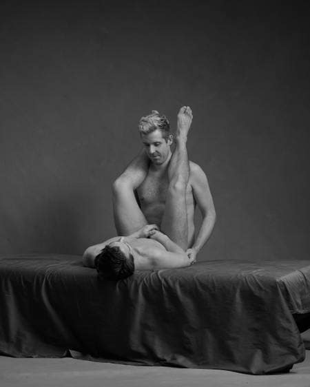 En naken mann ligger på ryggen på senga med begge bena rett opp og rundt halsen på en annen naken mann som står bak ham