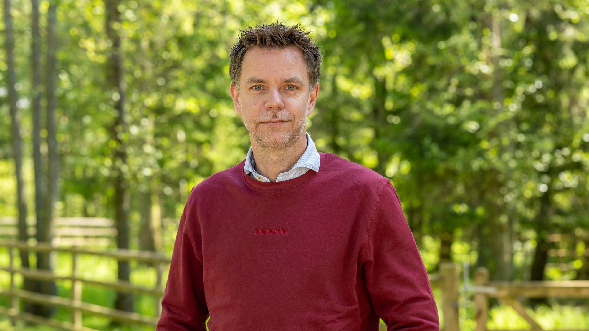 Mann med rød genser og kort grå/brunt hår ser inn i kamera. Han er omringet av grønn natur.