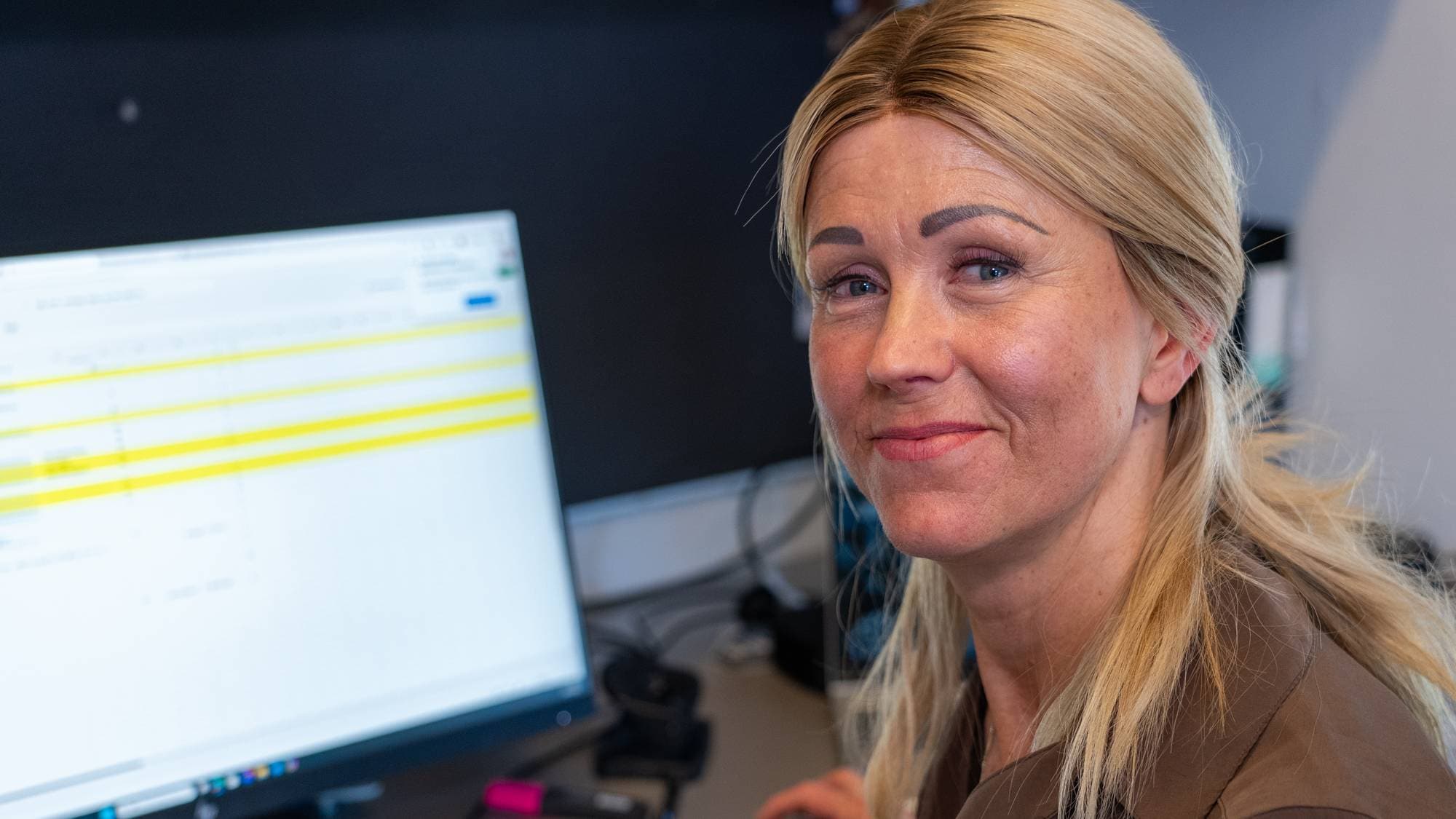 En kvinne som ser i kamera. Hun sitter foran en PC-skjerm.