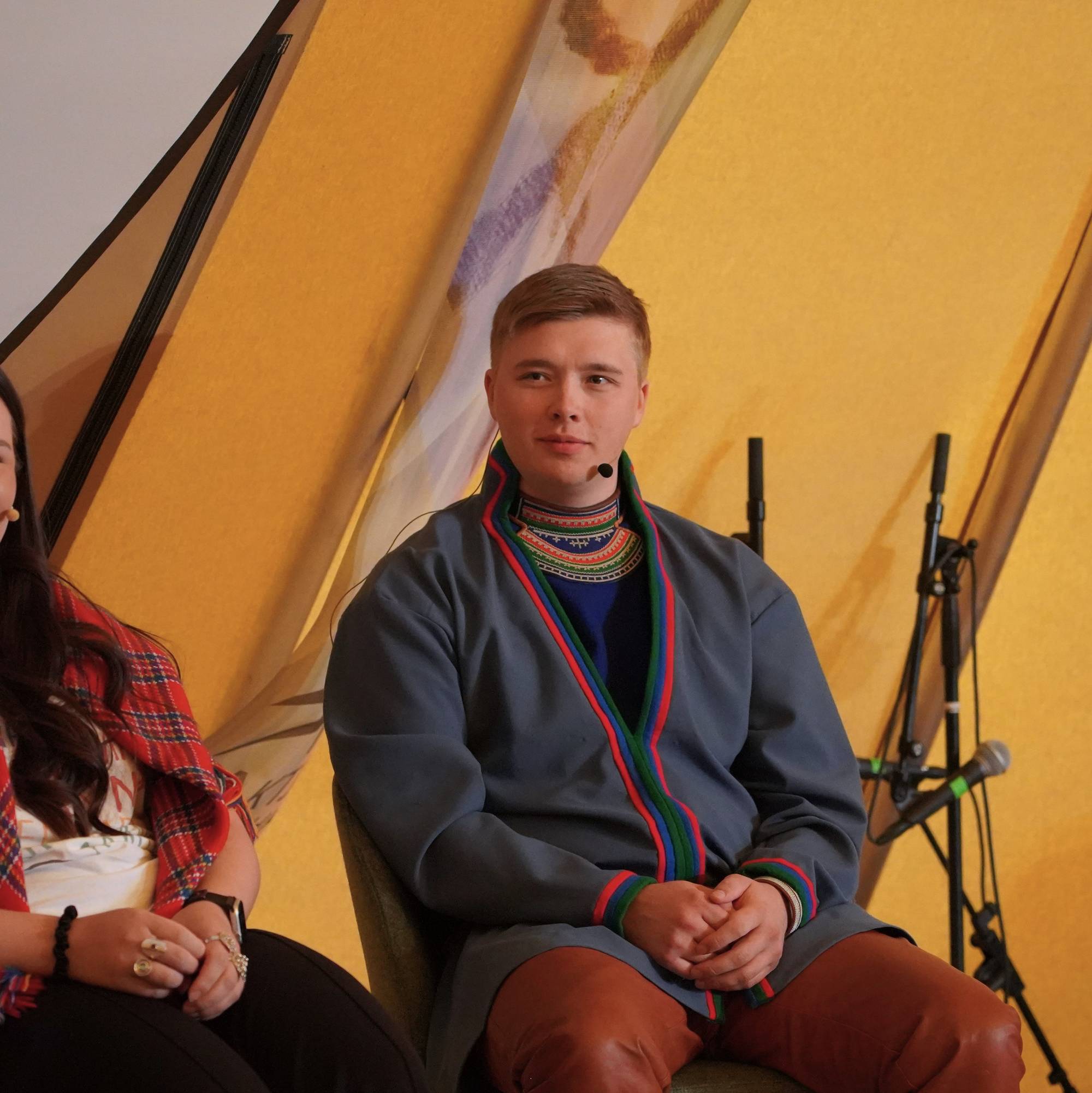 En kvinne i sjal og en mann i kofte sitter i et gult lavvo-telt under et arrangement. (Bildebeskrivelsen er laget av en KI-tjeneste)