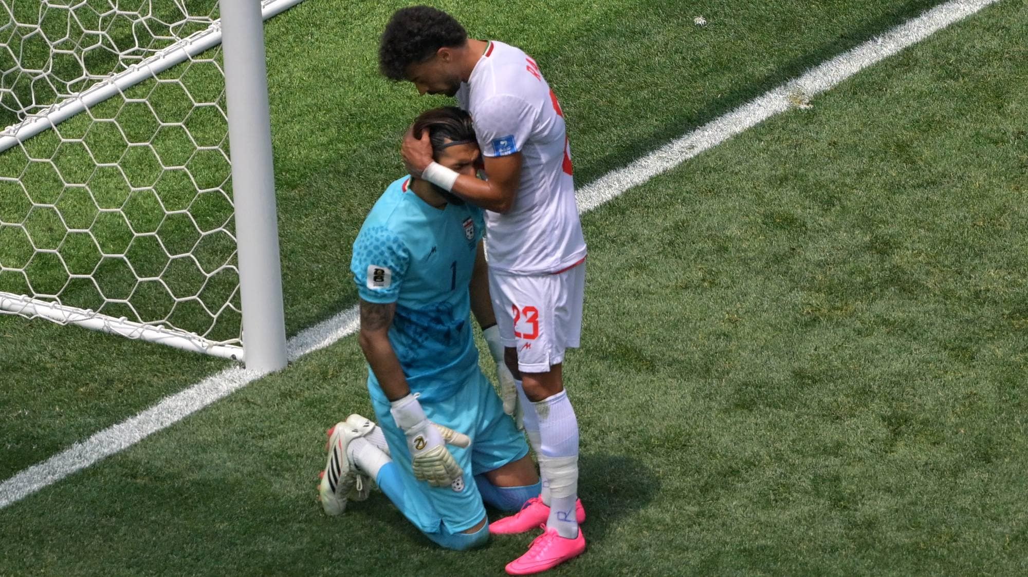 En fotballspiller i hvitt trøster en keeper i turkisgrønt som sitter på kne. Spilleren har en hånd på keeperens hode. Ved siden av er en fotballmål. Fotballbane med hvite linjer er synlig. (Bildebeskrivelsen er laget av en KI-tjeneste)