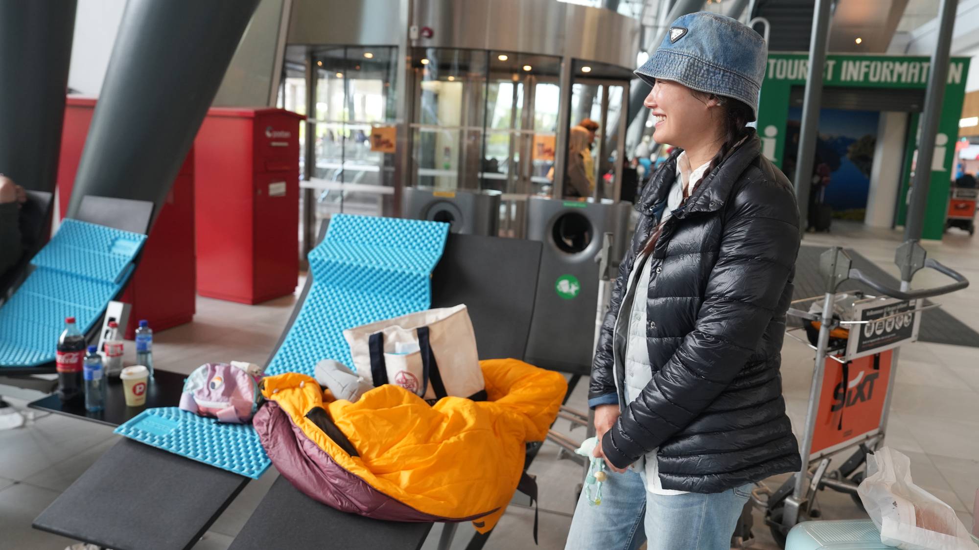 Kvinne med bøttehatt står ved sitteplasser på en flyplass med soveutstyr og bagasje. (Bildebeskrivelsen er laget av en KI-tjeneste)