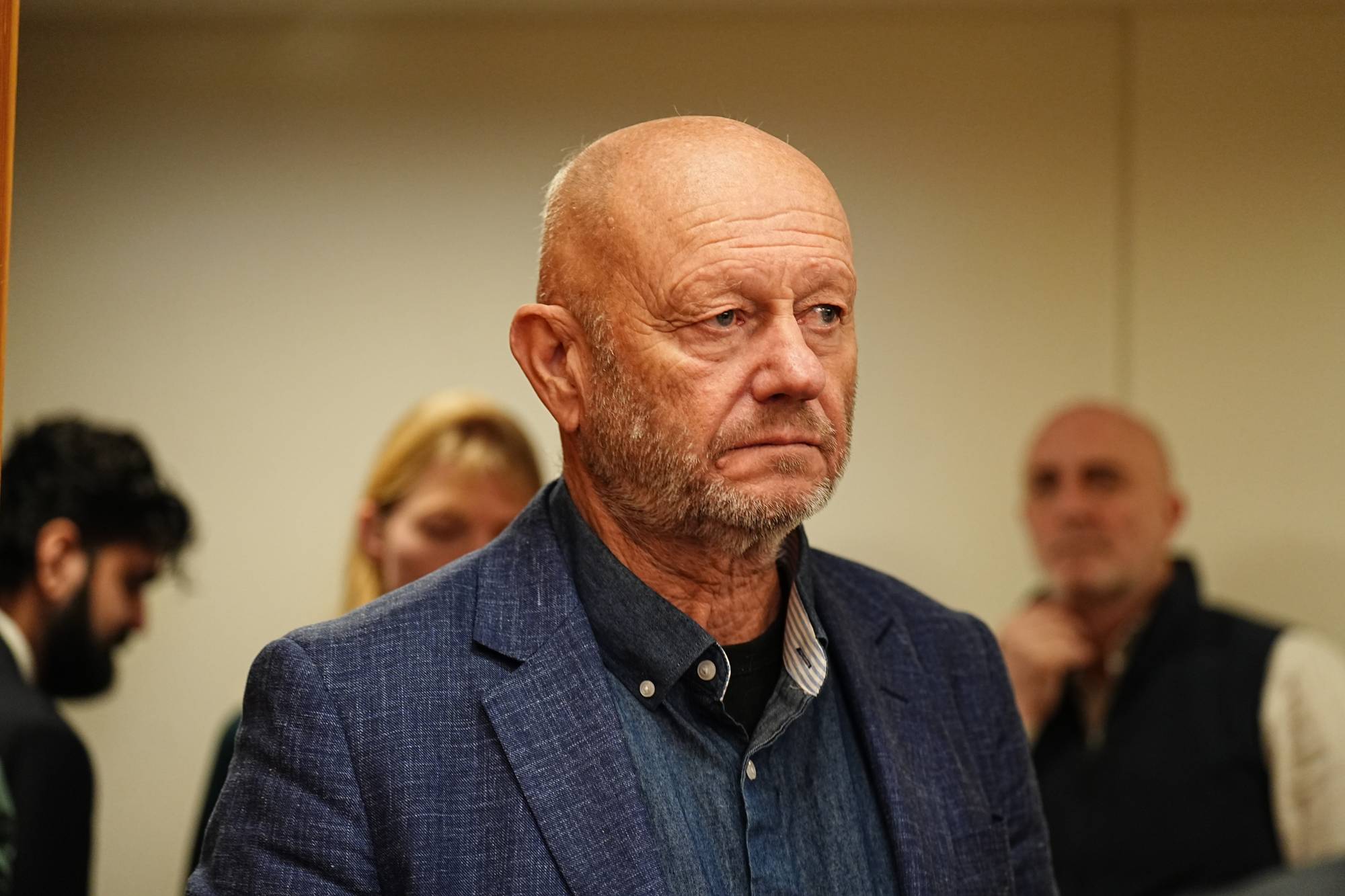 Et eldre menn står i en innendørs setting, iført en blå blazer og en mørk skjorte. Bak ham er flere personer delvis synlige, med fokus på ansikter som er delvis skjult. Omgivelsene er nøkterne, med en lys bakgrunn. Det er ingen åpenbare reklameelementer i bildet.