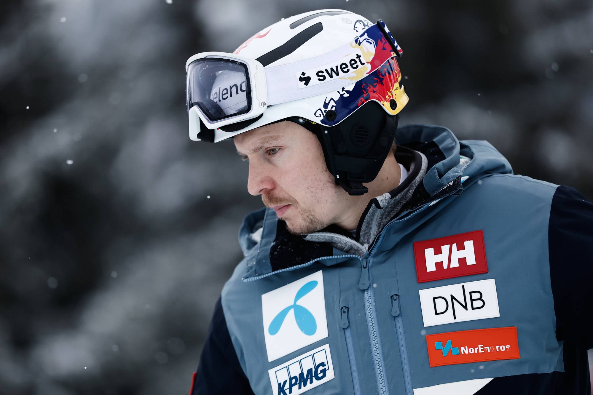 En skiløper i en blå jakke står utendørs i snøen, iført hjelm og briller. Det er snøfall, og bakgrunnen er uklar. Jakken har flere sponsorlogoer synlige, inkludert HH, DNB og KPMG. Skiløperens uttrykk og holdning tyder på konsentrasjon før konkurranse. (Bildebeskrivelsen er laget av en KI-tjeneste)