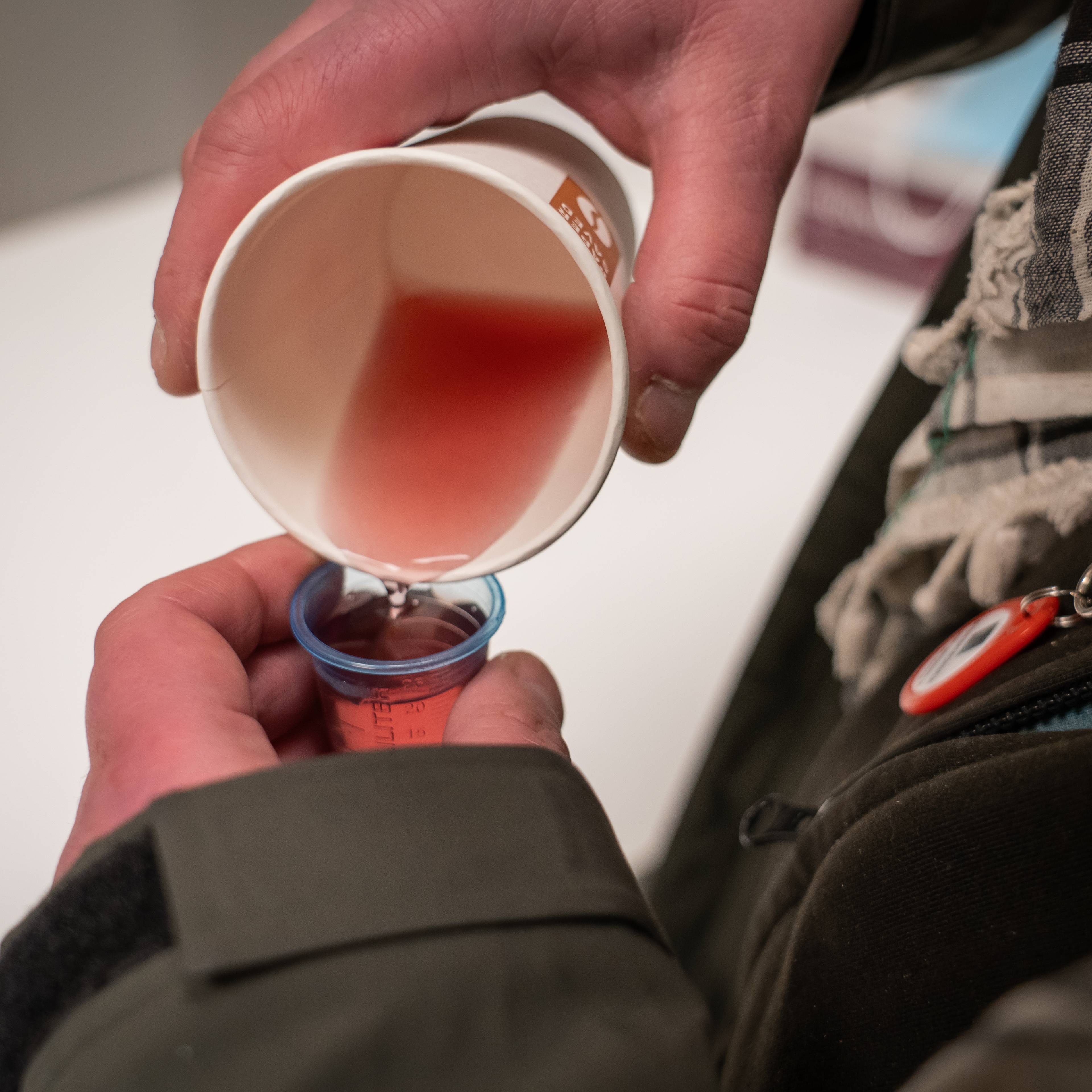 En person holder en papirbeholder med rød væske, som helles over i et målebeger. Hendene deres er synlige, og de er iført en jakke med en hette og en skulderstropp. Omgivelsene ser ut til å være innendørs, med en lys bakgrunn. (Bildebeskrivelsen er laget av en KI-tjeneste)