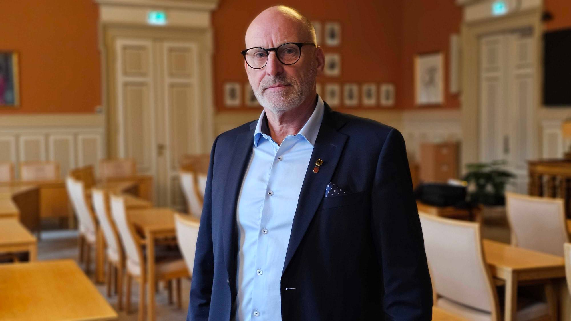 Eldre mann med briller og dress står i et møterom. (Bildebeskrivelsen er laget av en KI-tjeneste)