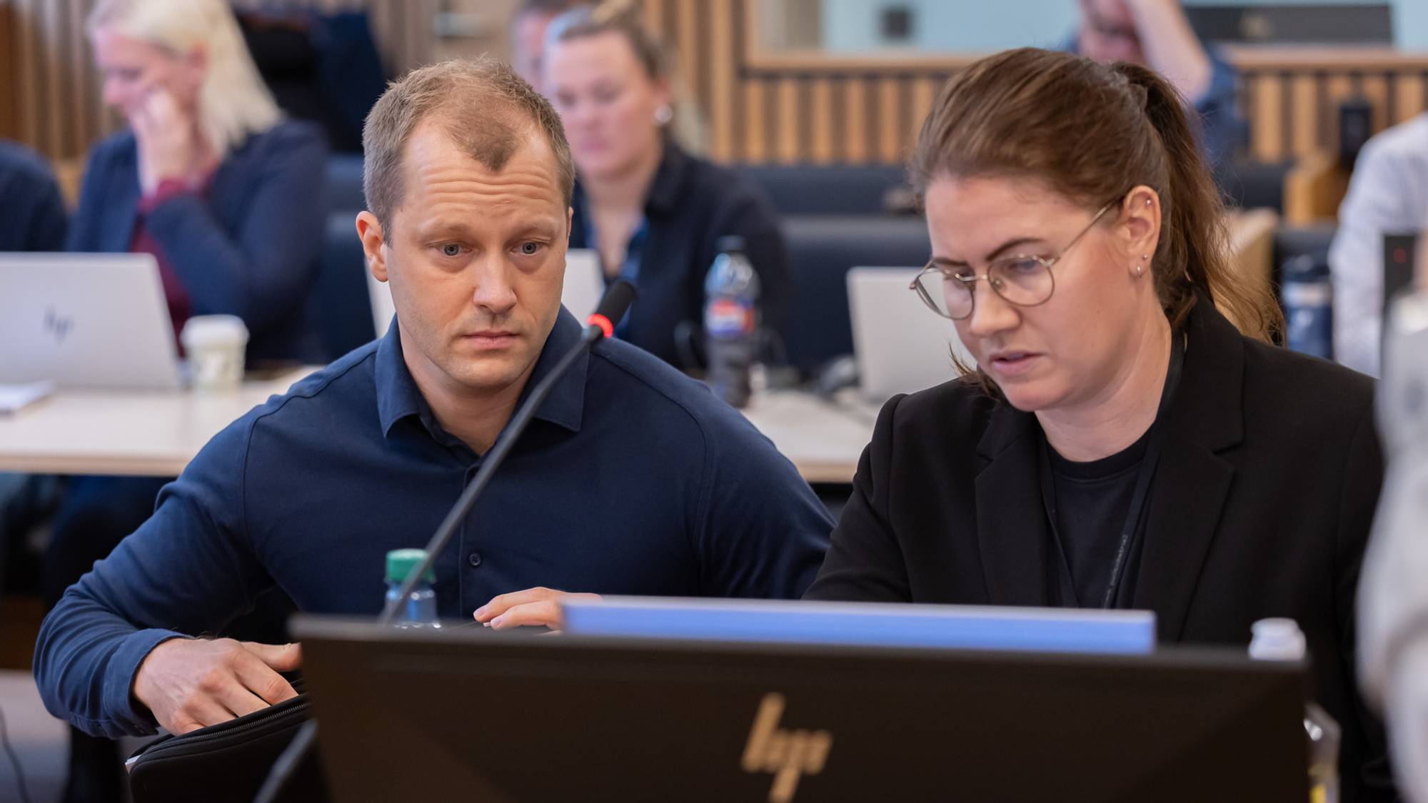 En mann (til venstre) og en kvinne (til høyre) sitter ved en vitnestol i en rettssal. De kikker på en PC. I bakgrunnen kan vi se noen folk.