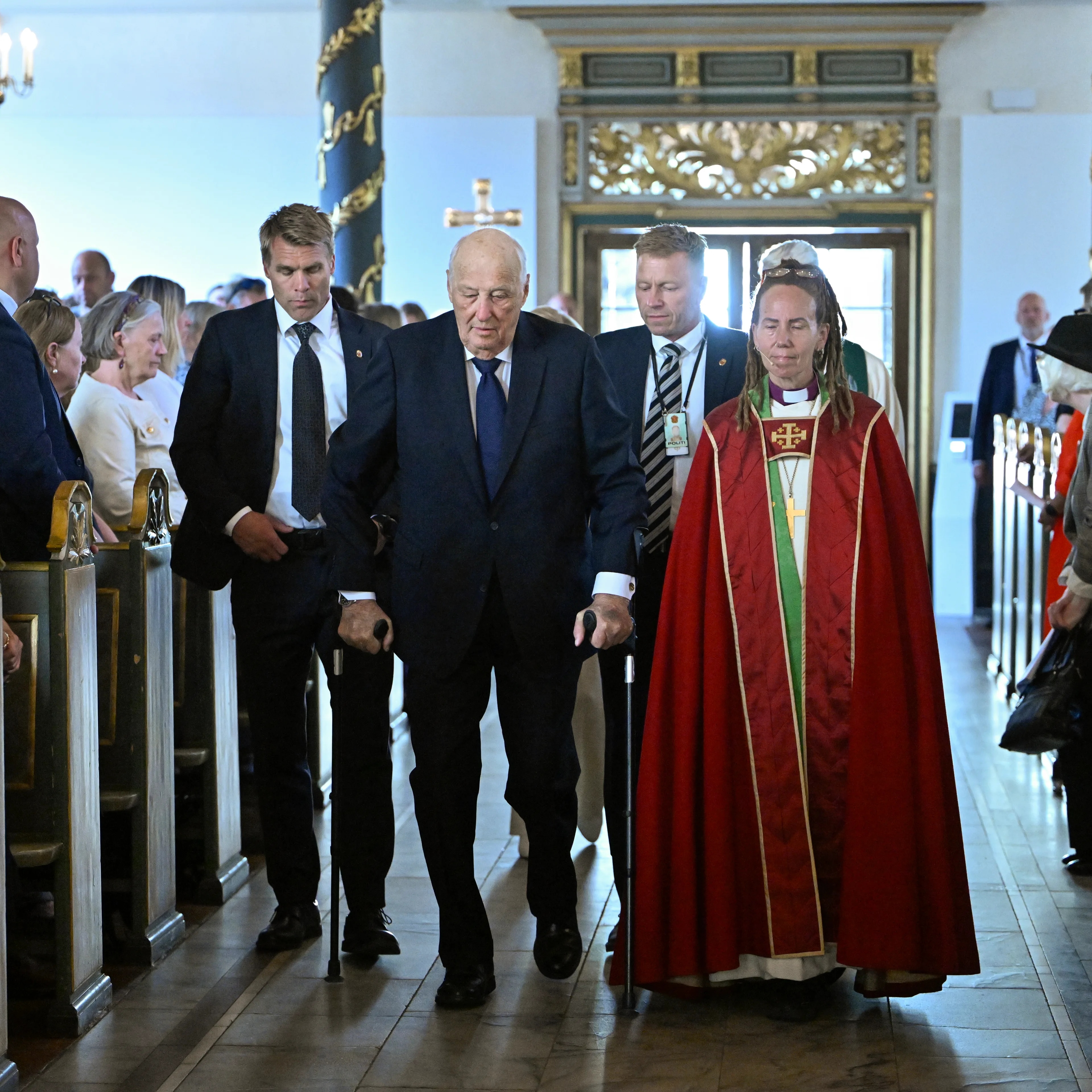 Kong Harald går opp kirkegulvet med støtte, flankert av en biskop og sikkerhetspersonell.
