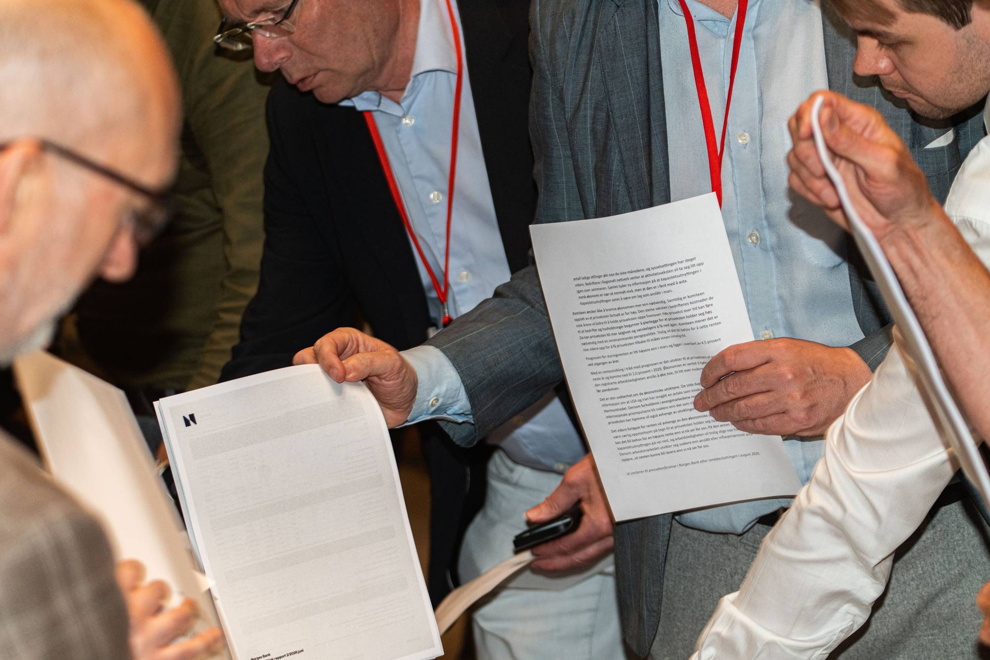 Flere menn ser på papirer under et møte. En av mennene holder frem et dokument foran seg. En annen mann med briller er i bakgrunnen. Dokumentet har et N-symbol og tekst. (Bildebeskrivelsen er laget av en KI-tjeneste)