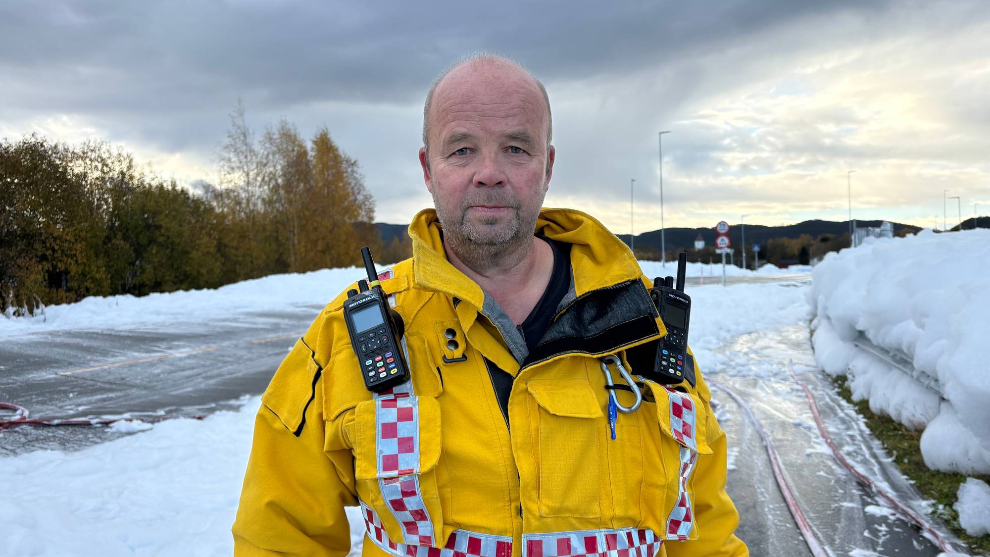 Brannmann i gul uniform med sambandsutstyr står utendørs ved en vei dekket av hvitt skum. (Bildebeskrivelsen er laget av en KI-tjeneste)