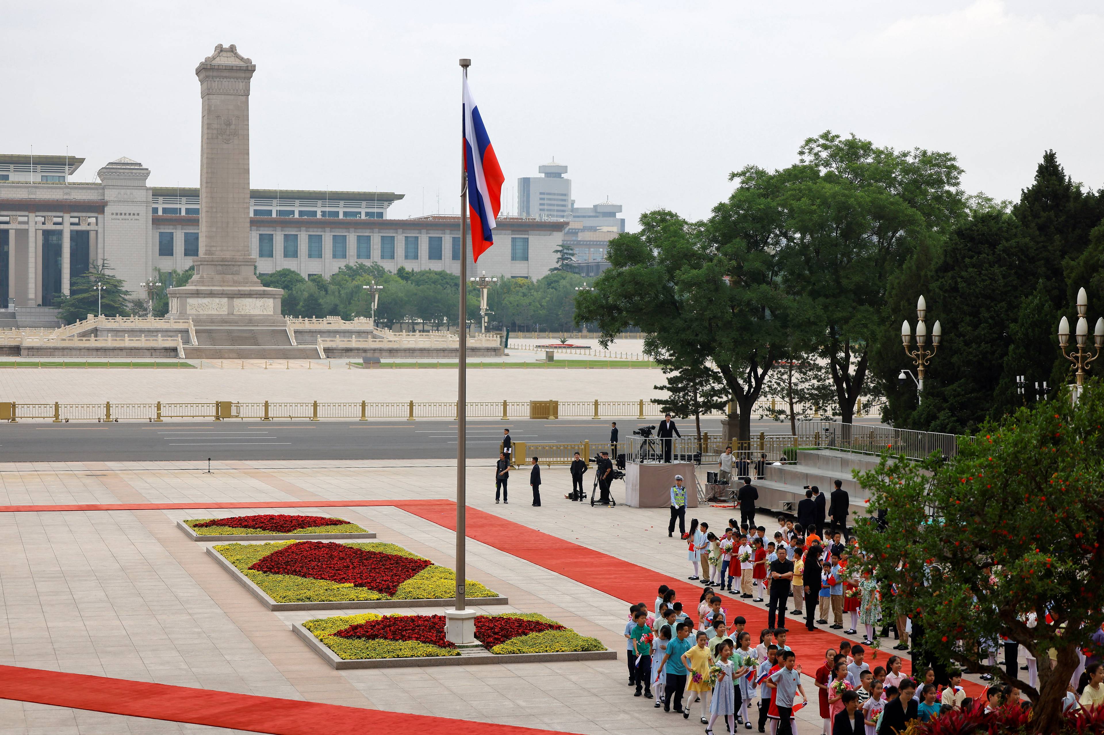 En russisk flagg vaier på en flaggstang foran en gruppe mennesker. I bakgrunnen er monumentet for Folkets helter og en stor bygning. En gruppe barn står i en rekke på en rød løper. (Bildebeskrivelsen er laget av en KI-tjeneste)