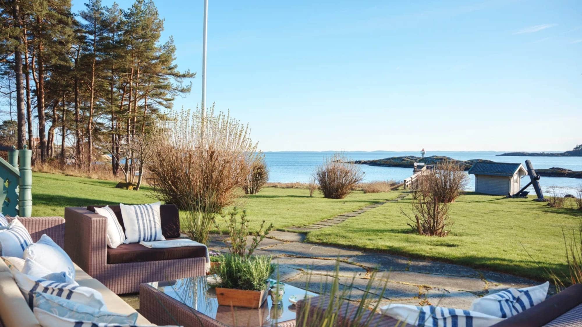 Utemøbler med puter står på en terrasse med utsikt over en gressplen og havet. I bakgrunnen er det trær og en liten bygning ved vannkanten. Solen skinner fra en klarblå himmel. (Bildebeskrivelsen er laget av en KI-tjeneste)