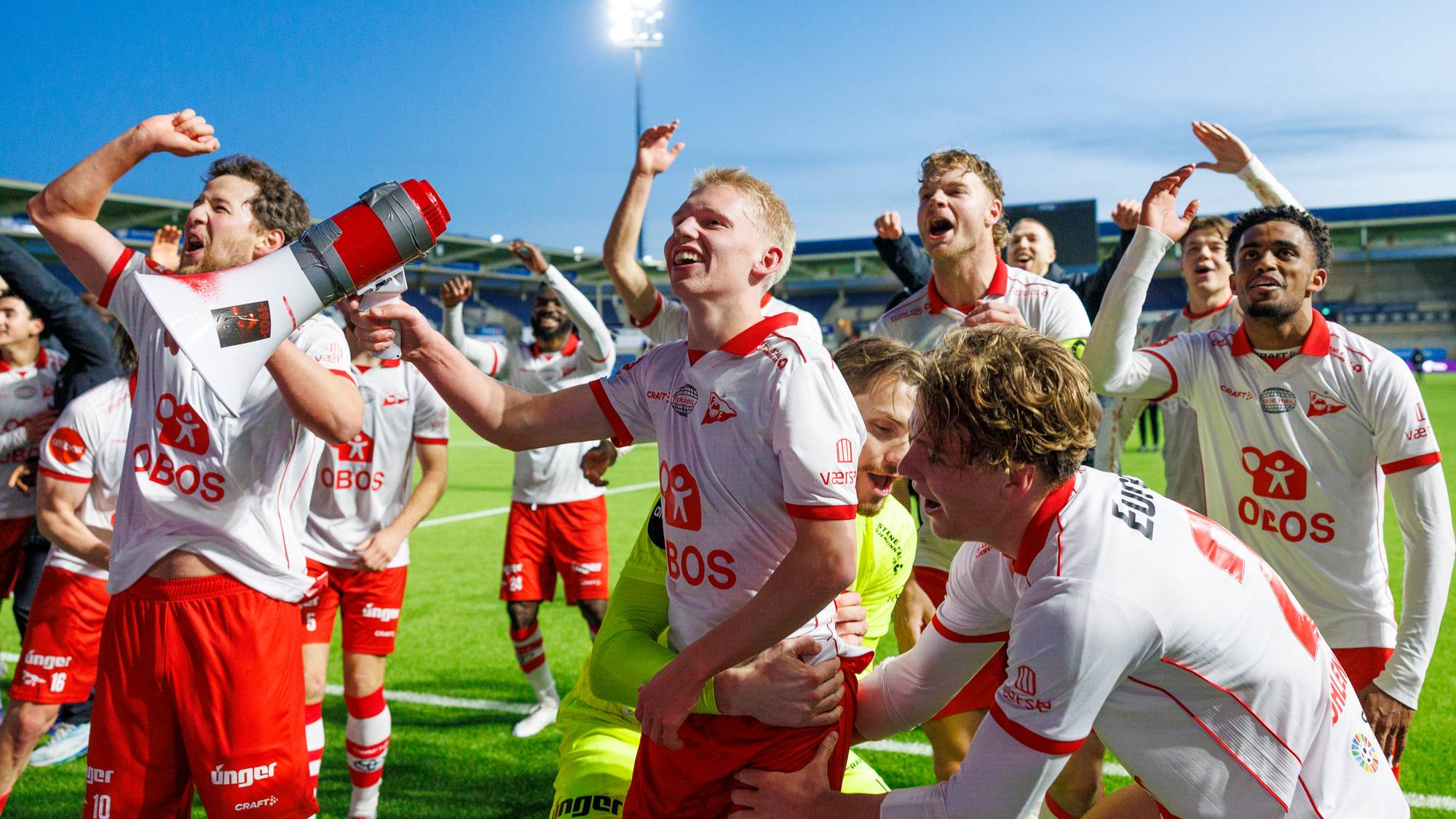 En gruppe fotballspillere i hvite og røde drakter feirer på en gressbane med et stadion i bakgrunnen. En spiller holder en megafon med påmalt bilde. Spillerne ser glade ut og gestikulerer mot himmelen. Det er kveld og stadionlysene er tent. (Bildebeskrivelsen er laget av en KI-tjeneste)
