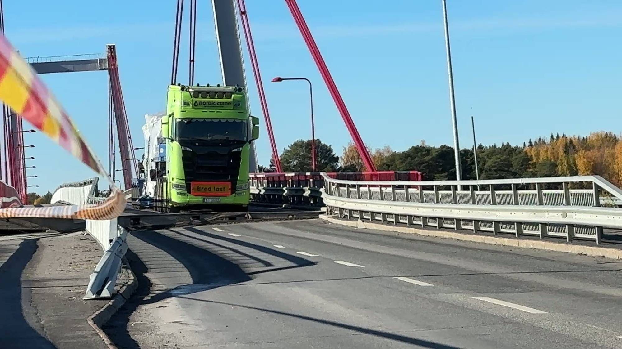 En grønn lastebil fra Nordic Crane med bred last kjører over en bro med røde stag. (Bildebeskrivelsen er laget av en KI-tjeneste)