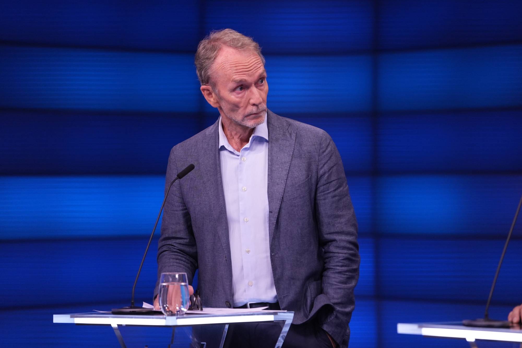 Mannen står ved en talerstol foran en blå bakgrunn. (Bildebeskrivelsen er laget av en KI-tjeneste)