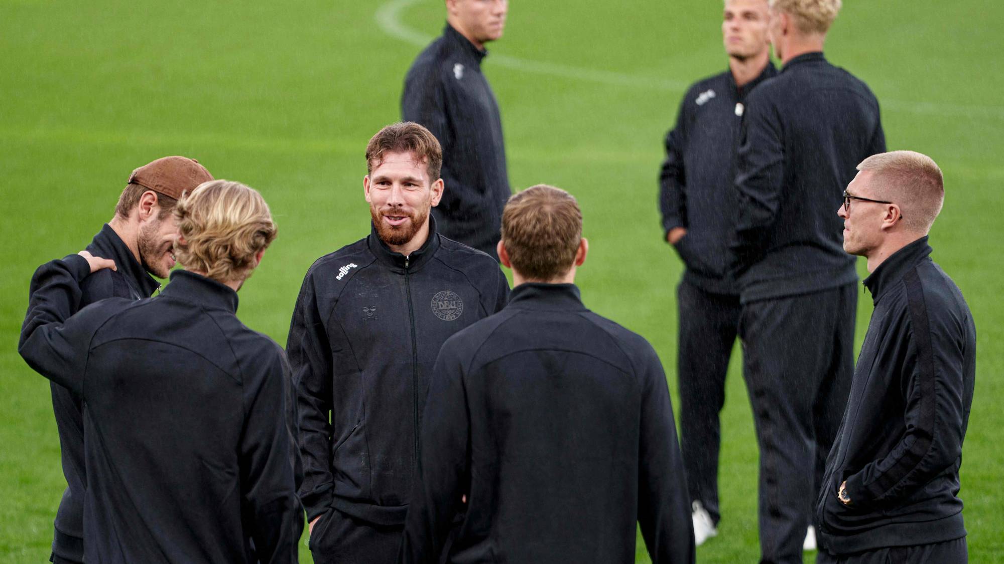 Fotballspillere i sorte treningsklær står i en samtale på en gressbane. (Bildebeskrivelsen er laget av en KI-tjeneste)
