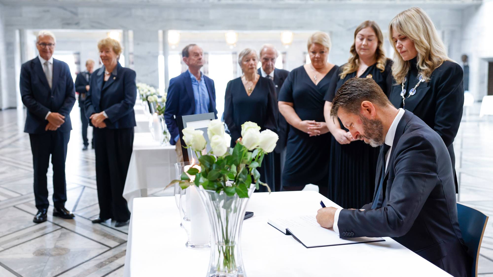 En mann signerer en protokoll med en gruppe mennesker stående bak seg i et marmorbygg. (Bildebeskrivelsen er laget av en KI-tjeneste)