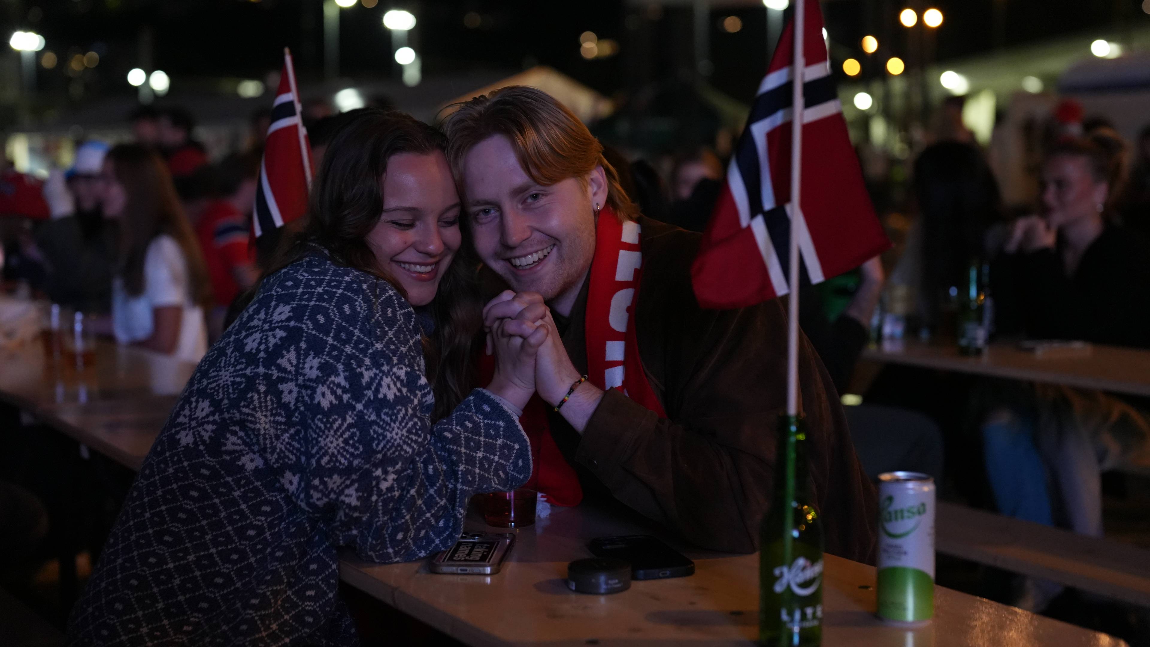 Et forelsket par holder hender og smiler til kameraet mens de sitter ved et bord. De har på seg et skjerf og en genser med norrøne mønstre. Et norsk flagg er stukket ned i en grønn flaske. Bildet er tatt utendørs om kvelden, med lys fra lyktestolper og andre lyskilder i bakgrunnen.