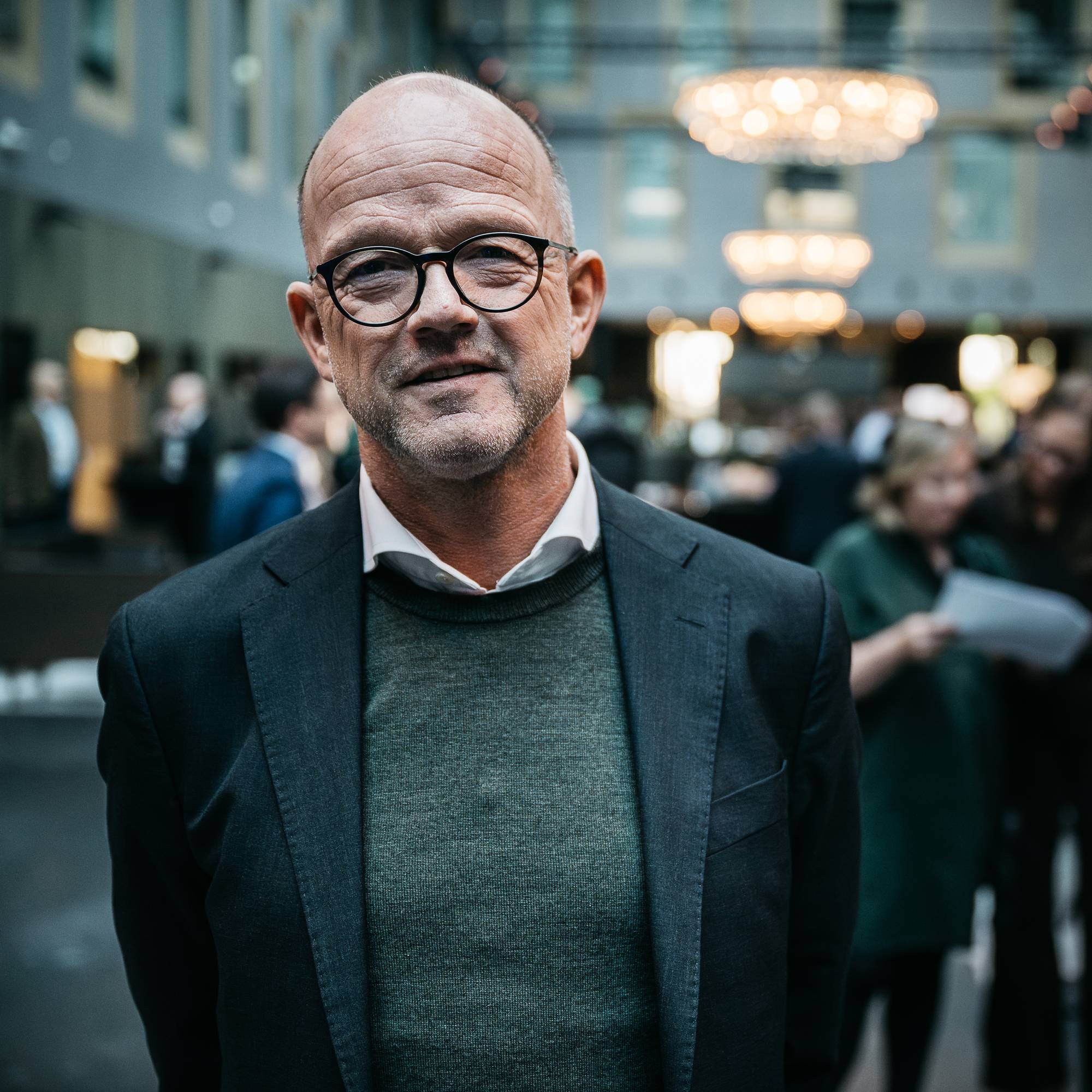 Bildet viser en mann iført dress og genser, som står i forkant av en folkemengde i en stor, moderne lokasjon. (Bildebeskrivelsen er laget av en KI-tjeneste)