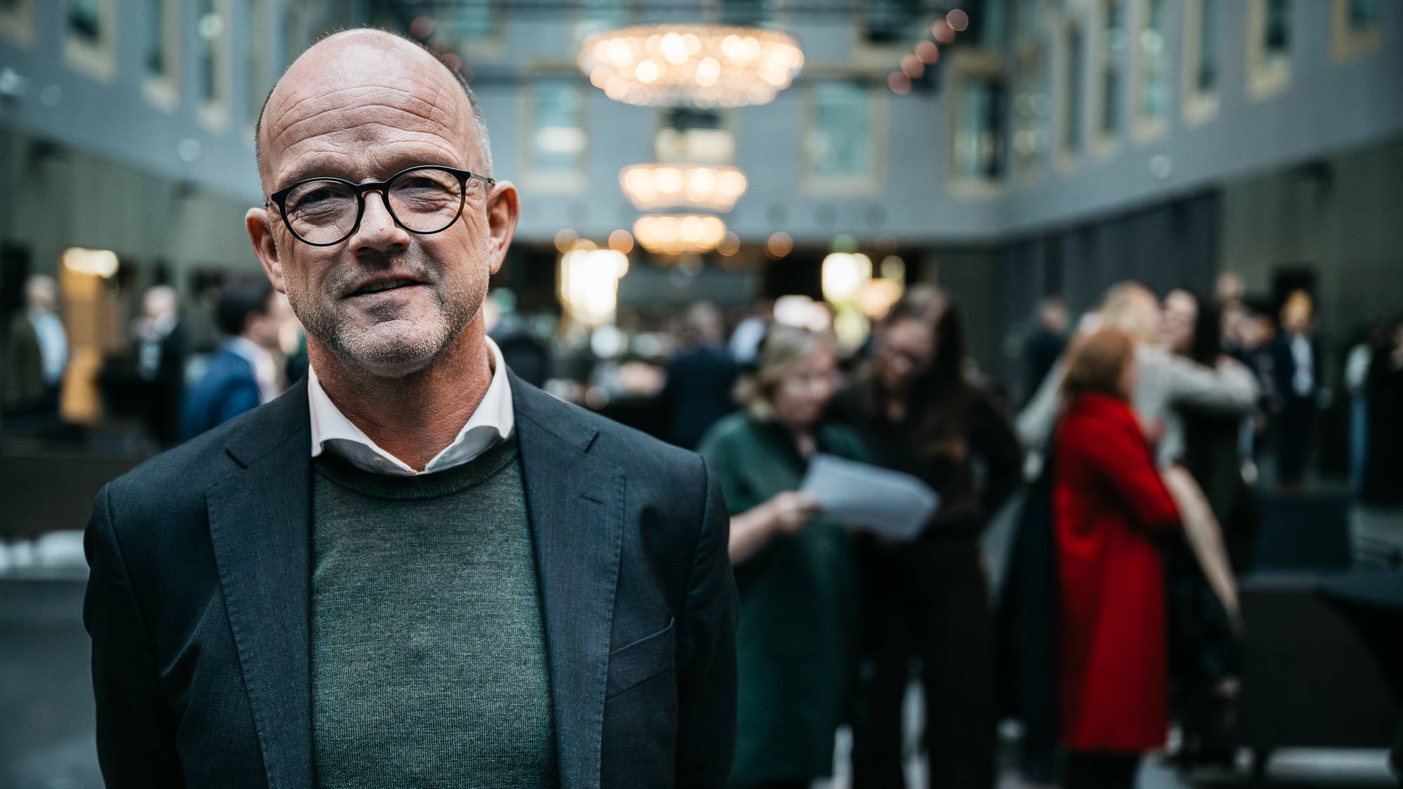 Bildet viser en mann iført dress og genser, som står i forkant av en folkemengde i en stor, moderne lokasjon. (Bildebeskrivelsen er laget av en KI-tjeneste)