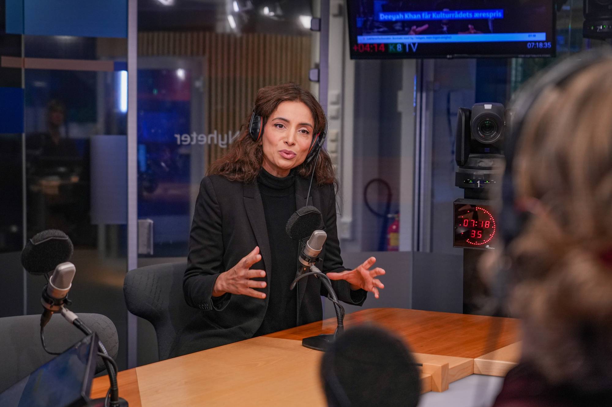Deeyah Khan sitter i et radiostudio og snakker inn i en mikrofon. (Bildebeskrivelsen er laget av en KI-tjeneste)