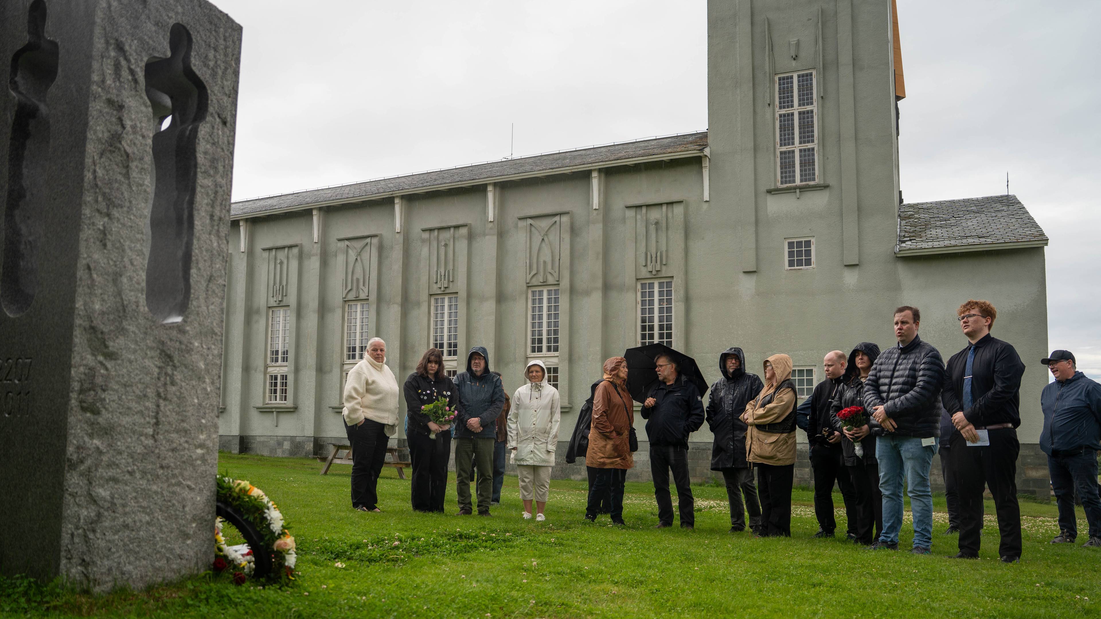 En gruppe mennesker står ved et minnesmerke utenfor en kirke. Noen holder blomster. (Bildebeskrivelsen er laget av en KI-tjeneste)