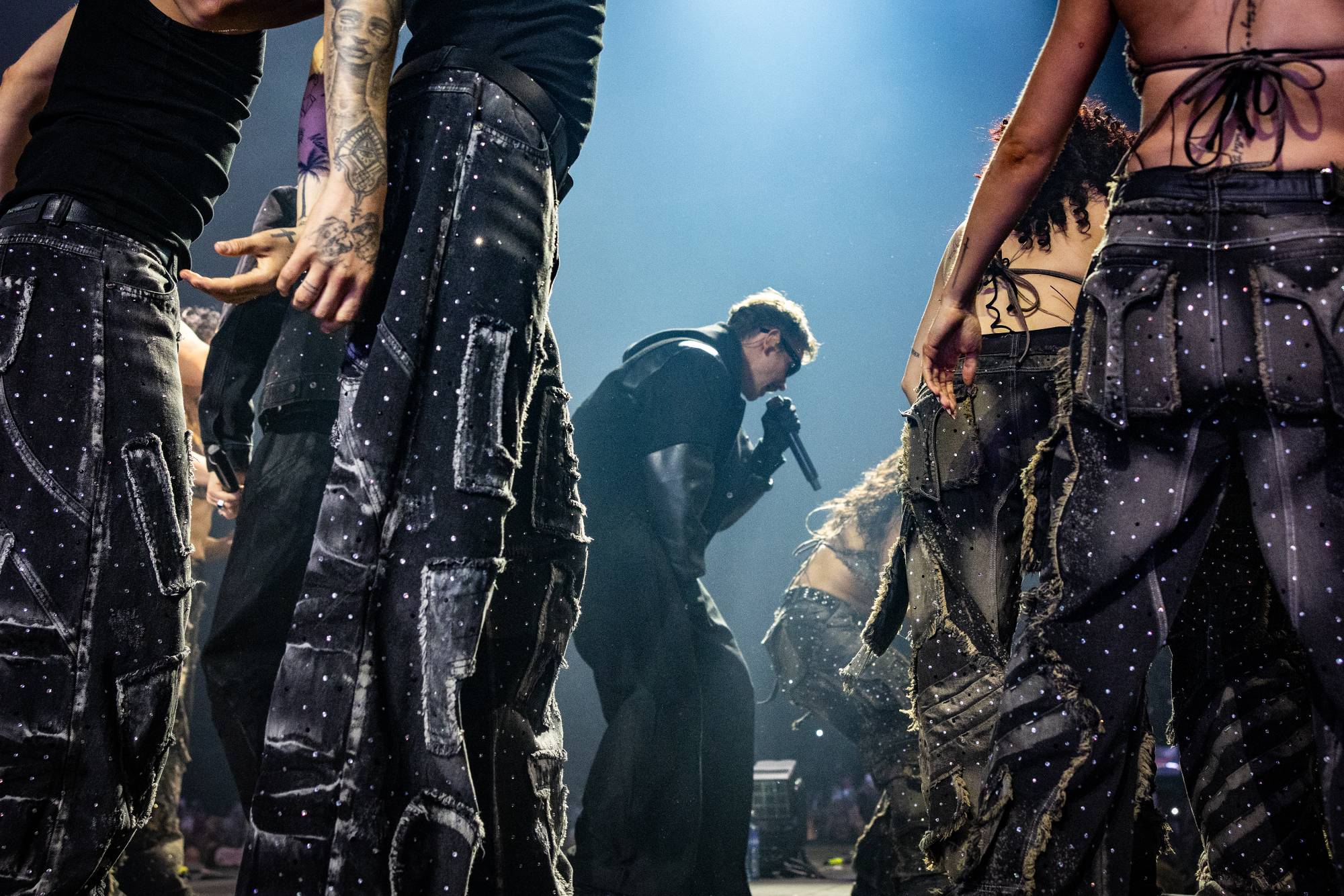 En artist synger i en mikrofon på en scene, med dansere rundt seg. Artisten har på seg en mørk jakke og briller. Danserne har på seg sorte bukser med glitterdetaljer. (Bildebeskrivelsen er laget av en KI-tjeneste)
