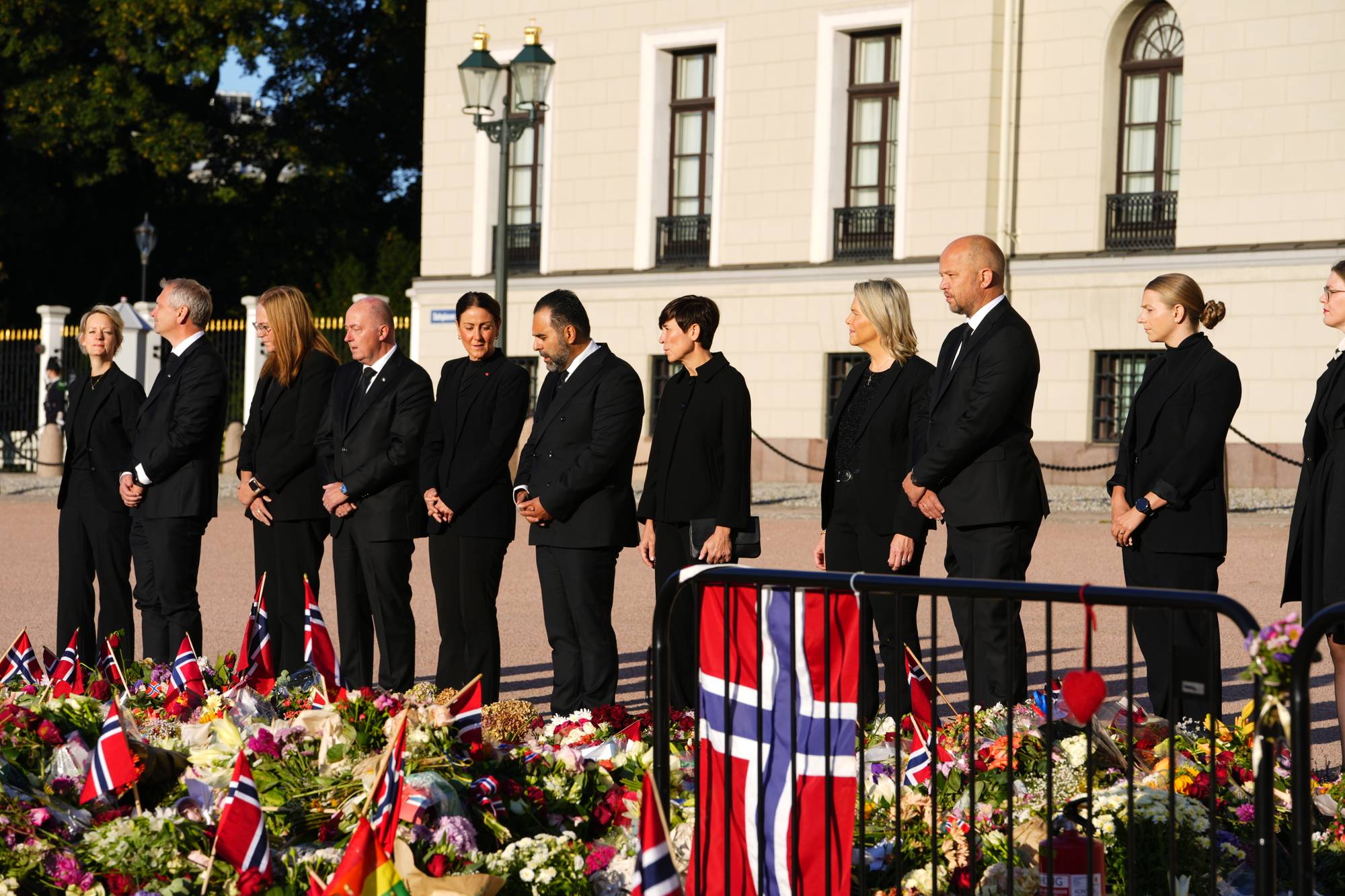 En rekke politikere fra Stortinget, kledt i sort står med hendene foldet foran seg og ser på blomsterhavet foran Slottet. I forgrunnen ser vi et norsk flagg og blomster. Solen skinner på slottets fasade. 