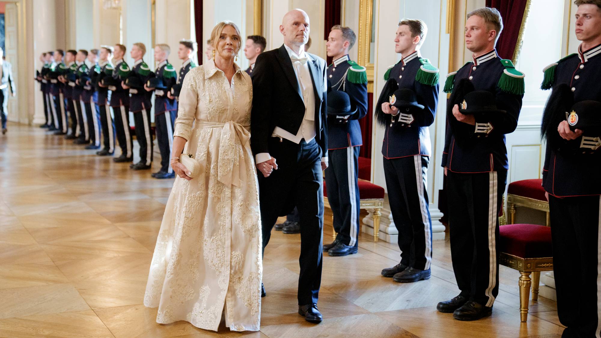En kvinne i lang, lys kjole og en mann i dress går forbi en rekke soldater. De andre soldatene står på rekke og rad, kledd i mørke uniformer med grønne detaljer. Rommet har store søyler og gulv med rutemønster. (Bildebeskrivelsen er laget av en KI-tjeneste)