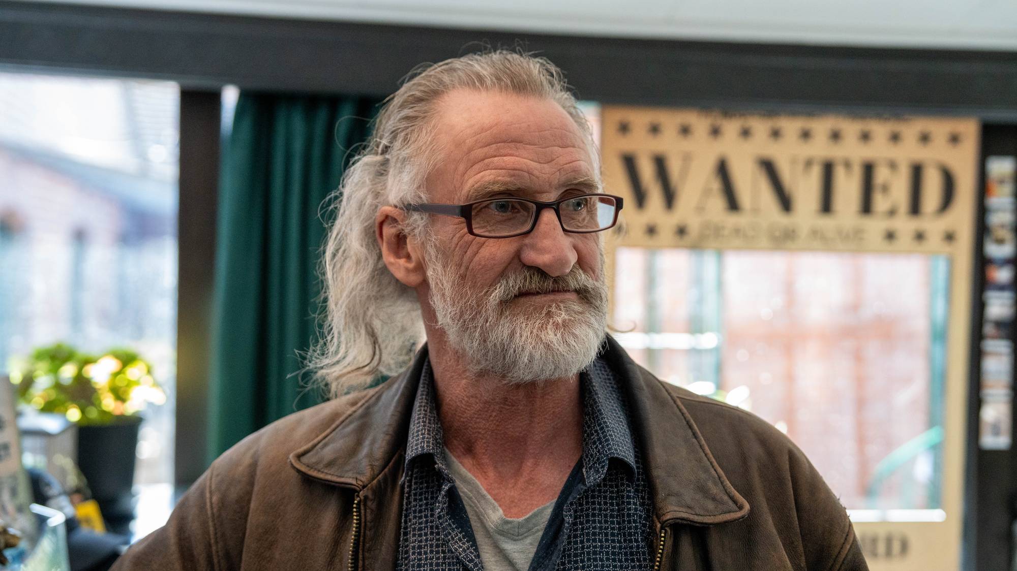 Et portrett av en eldre mann med briller og langt grått hår. Bak ham henger en plakat med teksten "WANTED". Mannen har på seg en brun skinnjakke over en rutete skjorte. (Bildebeskrivelsen er laget av en KI-tjeneste)