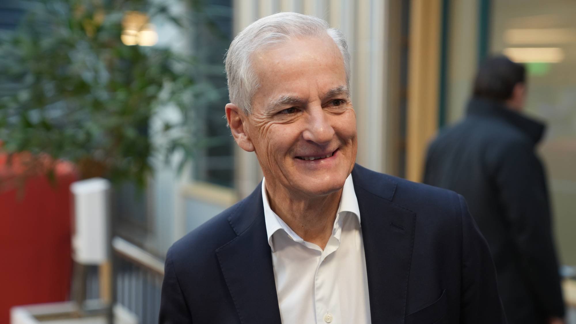 Jonas Gahr Støre, leder for Arbeiderpartiet, smiler til høyre. Han er kledd i mørk dress og hvit skjorte. I bakgrunnen skimtes en person i mørk jakke. Utenfor er grønne planter og en rød vegg. (Bildebeskrivelsen er laget av en KI-tjeneste)