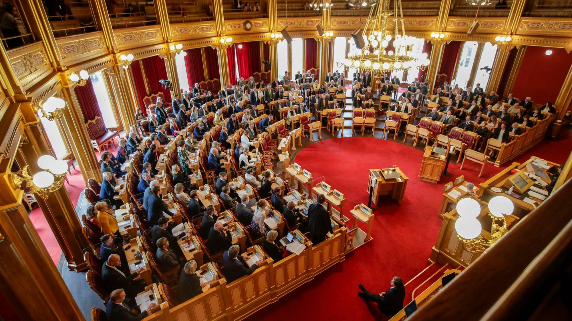 En gruppe mennesker sitter i en storsal med røde tepper og overdådige detaljer. De fleste ser ut til å være i en debatt eller møte med talerstoler og podier. Rommet har høye vinduer, store lysekroner og balkonger med flere tilskuere. (Bildebeskrivelsen er laget av en KI-tjeneste)