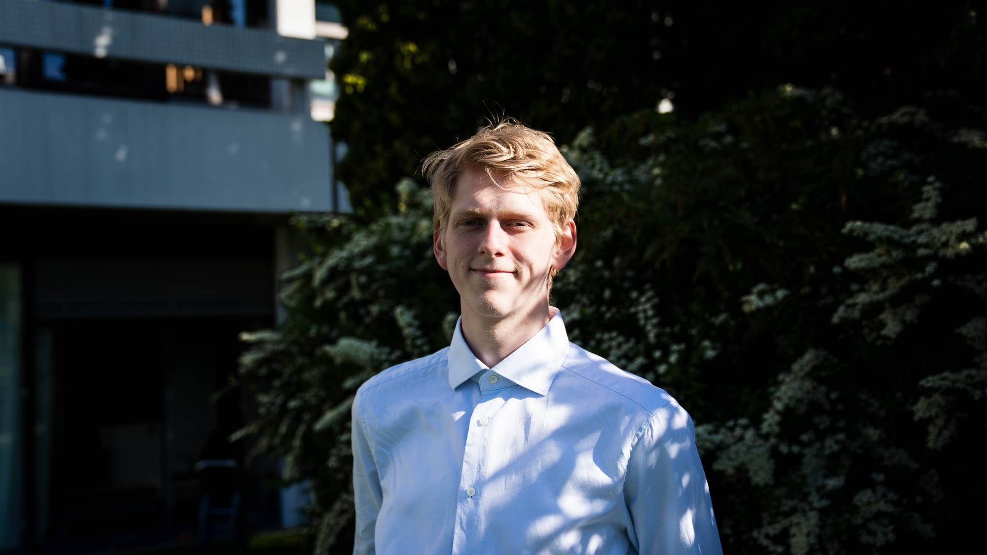 Portrett av en ung mann med blondt hår. Han står utendørs foran et tre og en bygning. Mannen har på seg en lys skjorte. Han smiler mot kameraet. 