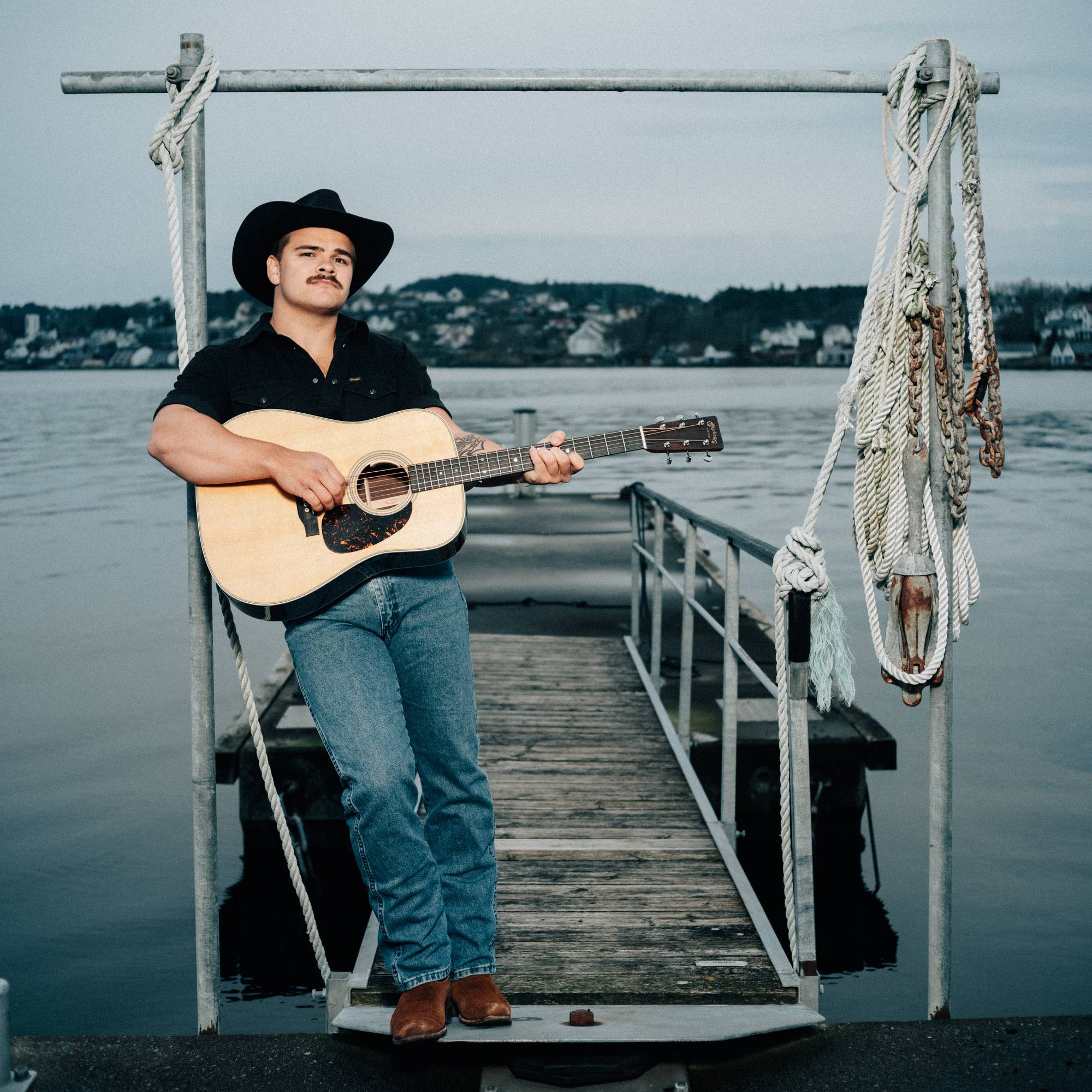 En mann med cowboyhatt og gitar står på en brygge ved vannet. Han har på seg en svart skjorte og blå jeans. Mannen ser mot kamera. (Bildebeskrivelsen er laget av en KI-tjeneste)