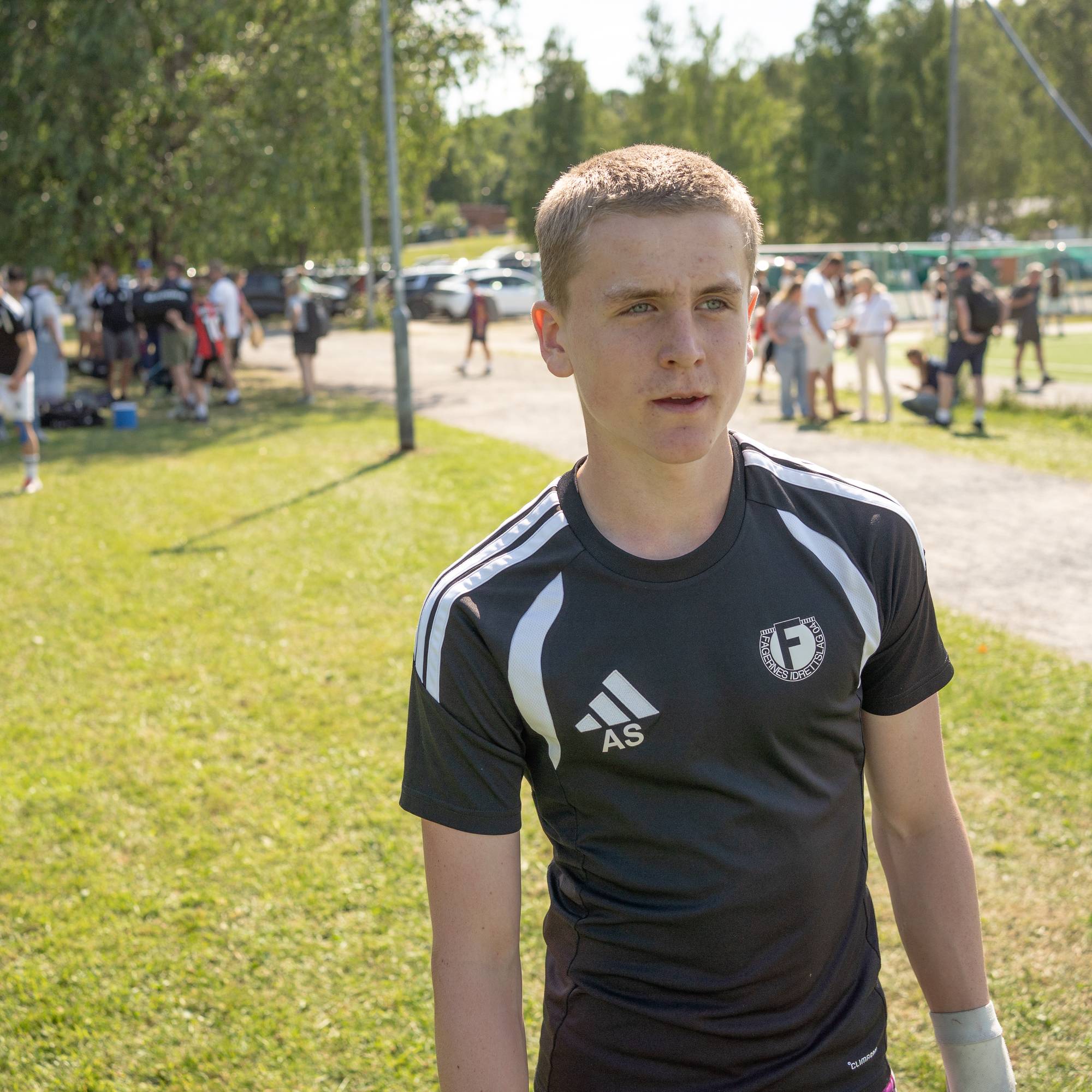 Ung mann i svart sportstrøye står ved en fotballbane med folk i bakgrunnen. (Bildebeskrivelsen er laget av en KI-tjeneste)