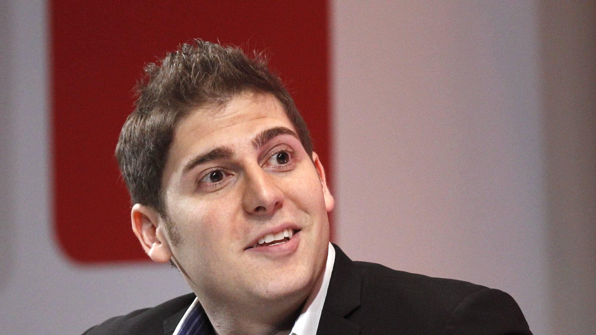 Eduardo Saverin ser til siden og smiler foran en rød og hvit bakgrunn. (Bildebeskrivelsen er laget av en KI-tjeneste)