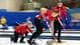 Lee Jin-man / AP / NTB Tre personer i røde jakker spiller curling på en isbane. En spiller bøyer seg ned mot en curlingstein, mens de andre står klare. Bakgrunnen viser deltakere i gult og blått. Hallen har blå vegger og en tilskuerrad. (Bildebeskrivelsen er laget av en KI-tjeneste)