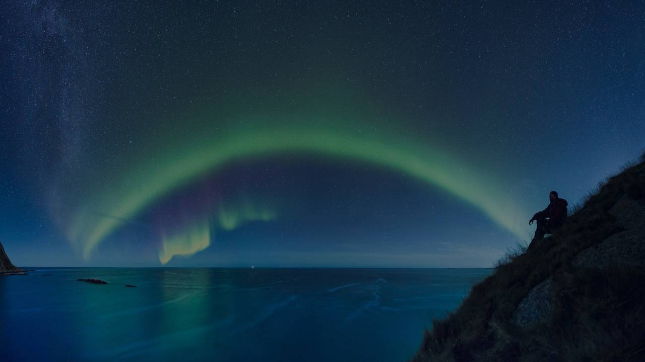 Person sittende på en klippe skuer utover havet på kveldstid. Nordlys spiller på himmelen.