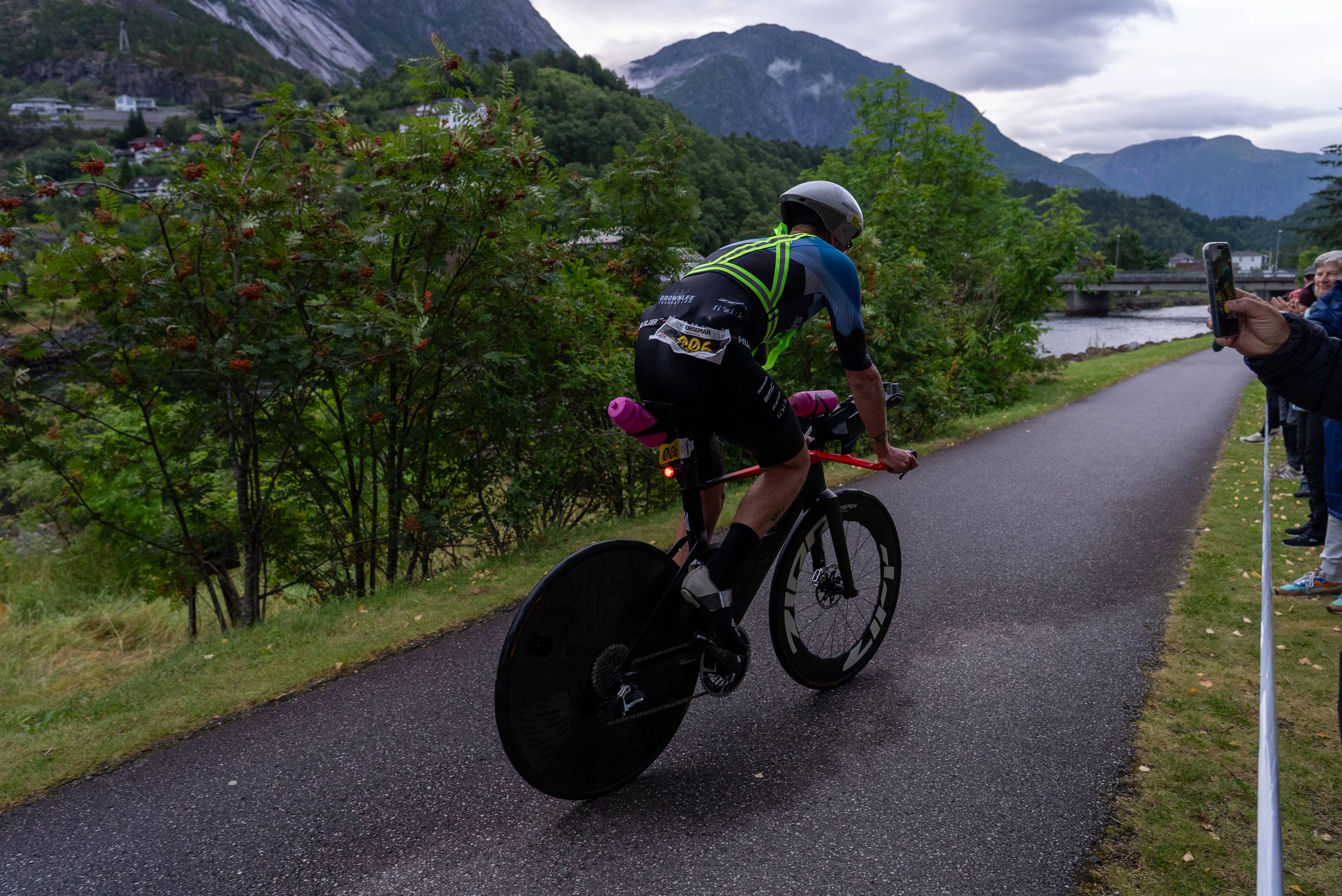 Syklist i triatlonkonkurranse sykler på en vei i et fjellandskap mens tilskuere ser på. (Bildebeskrivelsen er laget av en KI-tjeneste)