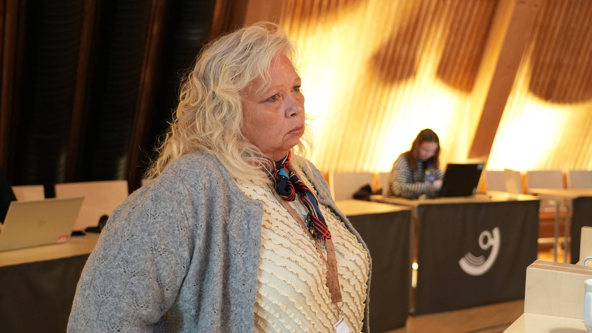 En kvinne med blondt hår står i et møterom med en kvinne med bærbar PC i bakgrunnen. (Bildebeskrivelsen er laget av en KI-tjeneste)